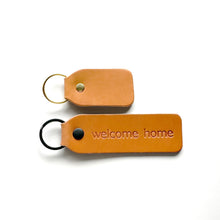 Two tan leather key tags on a white background, one personalized with 'welcome home' text and a black ring, the other plain with a gold ring.